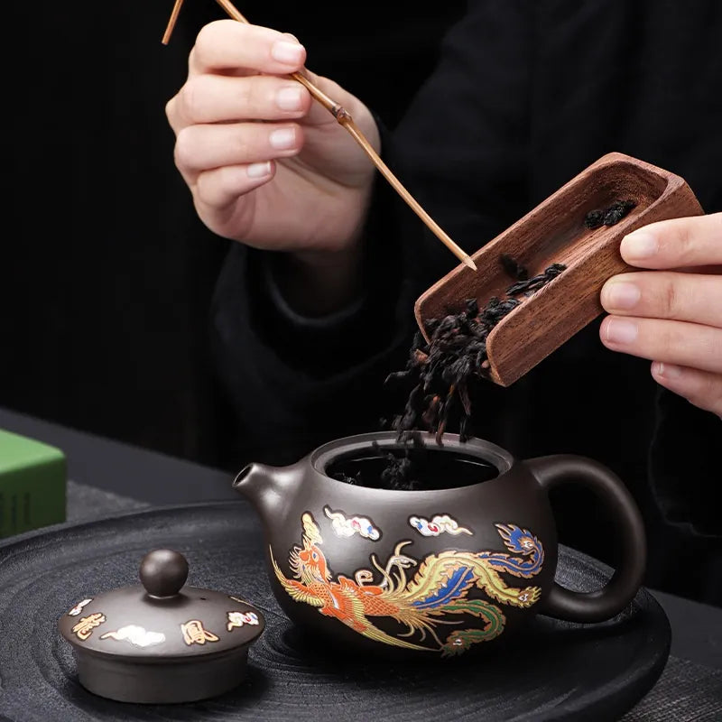 Person pouring tea leaves from a wooden scoop into a decorative teapot on a dark surface.